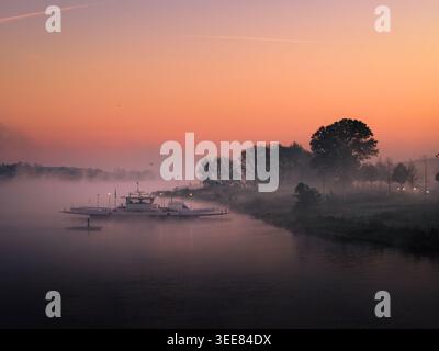 Aus der Vogelperspektive einer Fähre, die die nebelige Maas überquert, während eines leuchtenden orangen Sonnenaufgangs mit dickem Nebel über dem Wasser und Bäumen am Ufer in Kess Stockfoto