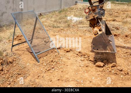 Eine Nahaufnahme des Metallschaufels eines Baggers, der sich in der Nähe eines tragbaren Siebs auf einer trockenen, staubigen Baustelle mit dünnem Gras befindet Stockfoto