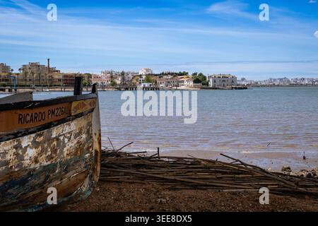 Ein rustikales, verlassenes Holzboot liegt am Strand an einer matschigen Küste mit einer entfernten Skyline der Stadt und ruhigem Wasser unter einem hellblauen Himmel Stockfoto