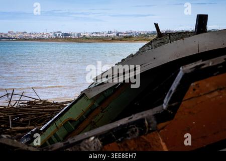 Nahaufnahme eines verrotteten Holzfischers, das an einem Ufer ruht, mit einer Küstenstadt, die über das Wasser sichtbar ist Stockfoto