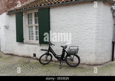 Schwarzes Fahrrad steht neben weißem Backsteinhaus mit grünen Fensterläden, gekacheltem Dach, Kopfsteinpflaster, ruhiger Straßenstimmung in Brügge. Fahrradkultur Stockfoto