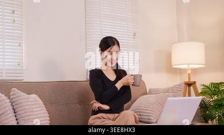 Asiatische Frau, die Kaffee genießt, während sie einen Laptop benutzt, bequem auf dem Sofa zu Hause sitzt und mit warmer Umgebungsbeleuchtung Stockfoto
