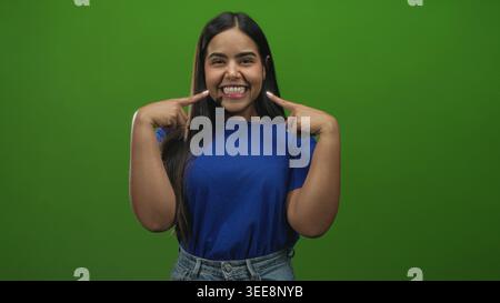 Die junge Frau zeigt mit den Fingern auf die Wangen und zeigt ein breites Lächeln, während sie ein blaues Hemd und eine Jeans auf einem grünen Bildschirm trägt; fröhlich verspielt. Stockfoto
