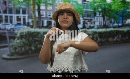 Die hispanische Frau zeigt auf der Straße Finger an Handgelenk, trägt Strohhut und weißes Hemd und wartet auf Ungeduld. Stockfoto