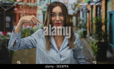 Junge hispanische Frau mit Brille zeigt beide Zeigefinger auf sich, während sie auf der belebten Straße lächelt; Selbstvertrauen. Stockfoto