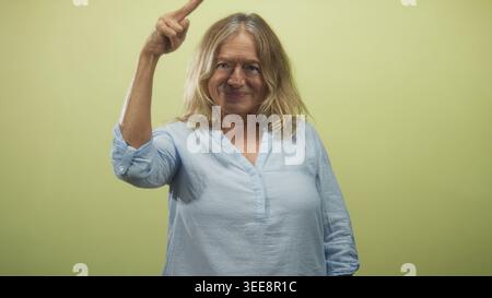 Die Seniorin zeigt in einem Studio vor einem blassgrünen Hintergrund mit dem Finger auf den Tempel, lächelt und entspannt; ruhiges Selbstvertrauen. Stockfoto