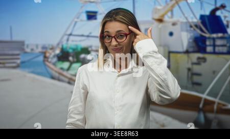 Die Frau zeigt mit dem Finger auf den Tempel am Straßenpier mit vertäuten Booten und Dock, trägt rote Brille und weißes Hemd; durchdachte Idee. Stockfoto