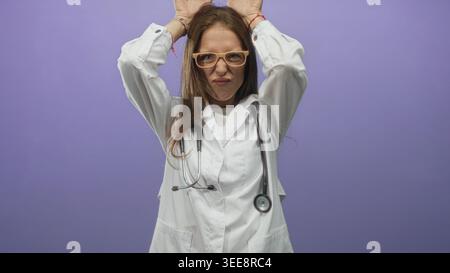Junge hispanische Ärztin mit weißem Mantel und Brille mit Stethoskop und Händen am Kopf im lila Atelier; Frustration. Stockfoto