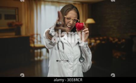 Frau Ärztin mit Stethoskop zeigt Finger auf Kamera in einem Wohnzimmergebäude, während sie rotes Herzspielzeug hält; fördert die Herzgesundheit. Stockfoto