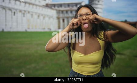 Frau zieht die Wangen und streckt die Zunge vor dem Gebäude aus, trägt gelbe Decke und Brille; verspielter Unfug. Stockfoto