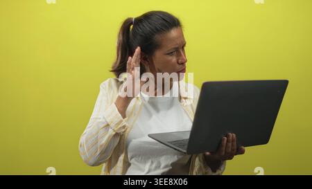 Eine brünette Frau mittleren Alters hält mit einer Hand einen Laptop und schränkt ihr Ohr mit der anderen, während sie im Studio mit gelbem Hintergrund auf den Bildschirm schwindelt Stockfoto