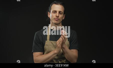 Mann mit langen Haaren, die Hände klatschen, im Studio mit schwarzem Hintergrund, Schürze und schwarzem T-Shirt; Besinnung. Stockfoto