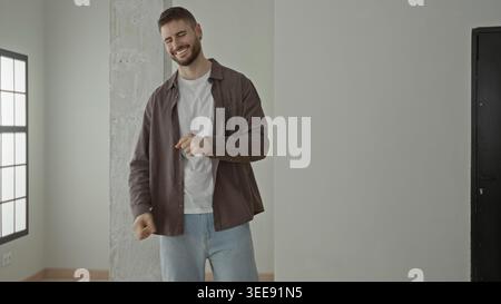 Mann klatscht mit den Händen und lächelt, während er sich in einem hellen Studio mit Säule und Fenster bewegt; Freude feiern. Stockfoto