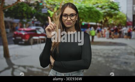 Junge Frau zeigt den Zeigefinger nach oben auf die Straße, Arme gefaltet, Brille und Jeans getragen; Vertrauensunterweisung. Stockfoto