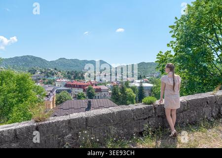 Frau in einem Blumenkleid, das an einer historischen Mauer steht und den malerischen Blick auf die Stadt Cetinje genießt. Friedliche morgendliche Atmosphäre, Montenegro Stockfoto