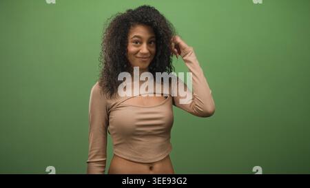 Eine junge Frau mit lockigen Haaren zeigt den Finger mit einem wissenden Lächeln vor grünem Studio-Hintergrund nach oben; verspielte Neugier. Stockfoto