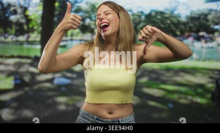 Frau in einem gelben, abgeschnittenen Tanktop zeigt die Daumen hoch und Daumen nach unten Gesten mit ausdrucksstarkem Gesicht in einem Studio-Set, das einem Park ähnelt; Unentschlossenheit Zweifel. Stockfoto