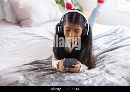 Indisches Mädchen im Schulalter, das auf dem Bett liegt, einen Pullover trägt, das Telefon hält und Kopfhörer hört Stockfoto