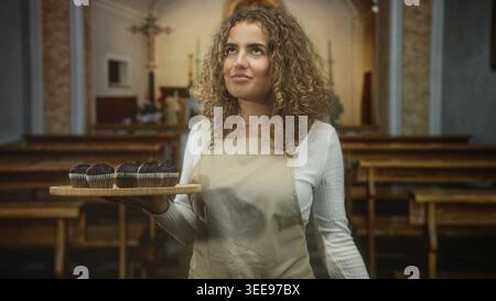 Frau, die ein Holztablett mit Schokoladenmuffins hält, Hand sichtbar, in einem Kirchengebäude; Heiterkeit. Stockfoto