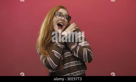 Rothaarige Frau, die Hände an die Brust schließt, lächelt mit geschlossenen Augen vor rosa Hintergrund in einem Studio; Freude. Stockfoto