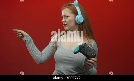 Junge rothaarige Frau, die mit dem Finger zeigt, während sie ein blaues Gamepad hält und Katzenohrkopfhörer in einem Studio mit rotem Hintergrund trägt; Frustrationsherausforderung. Stockfoto