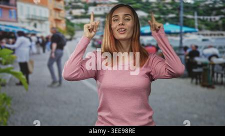 Frau zeigt beide Zeigefinger nach oben und zeigt Finger und Handflächen auf einer belebten Straßenpromenade; hoffe Neugier. Stockfoto