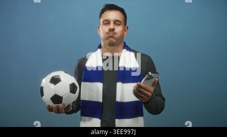 Mann, der Fußball und Smartphone hält, die Lippen und die Augen mit einem blauen und weißen Schal im Studio verschlossen hat; Freude an der Begeisterung der Fans. Stockfoto