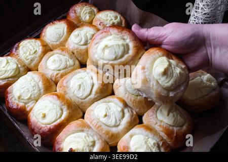Ein Tablett mit goldenem, hausgemachtem Gebäck, garniert mit cremigen weißen Strudeln, wahrscheinlich Käse oder Pudding. Eine Hand nimmt ein weiches, frisch gebackenes Brötchen auf. Stockfoto