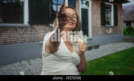 Die hispanische Seniorin zeigt mit dem Finger und hält 20-Dollar-Banknoten vor einem Gebäude; Selbstüberzeugung. Stockfoto