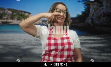 Hispanische Seniorin mit grauen Haaren und Brille, die eine rote karierte Schürze trägt, zeigt den Finger an die Nase und lächelt vor einem Studio-Strand-Hintergrund; fröhlich. Stockfoto