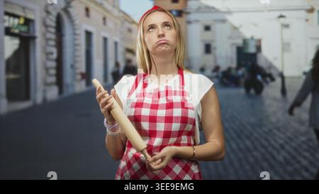 Chefkoch in roter Gingham-Schürze, die eine hölzerne Nudelnadel hält und mit offenem Unterarm auf einer kopfsteingepflasterten Straße zuckt; Frustration. Stockfoto