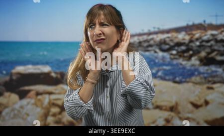 Frau, die das Ohr mit der Hand schröpft und sich nach vorne beugt, mit gestreiftem Hemd und Creolen im Studio Seaside Rocks; Verwirrung beim Hören. Stockfoto