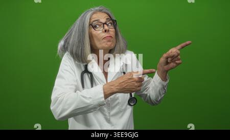Frau Ärztin mittleren Alters grauhaarig mit weißem Mantel und Stethoskop zeigt Finger nach rechts im grünen Atelier; Skepsis. Stockfoto