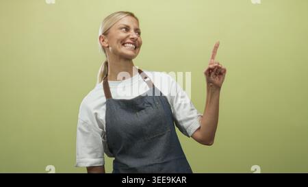 Frau aus Denim-Schürze und weißem T-Shirt zeigt den Zeigefinger nach oben und lächelt im Studio; fröhliches Selbstvertrauen. Stockfoto