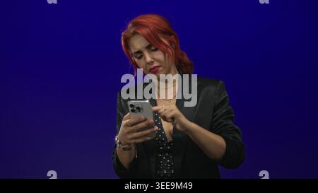 Junge hispanische Frau, die im Studio vor tiefviolettem Hintergrund mit dem Zeigefinger auf das Smartphone klopft; frustrierendes technisches Problem. Stockfoto
