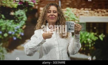 Die hispanische Frau im mittleren Alter zeigt mit dem Finger auf den Becher und liest: ich bin der Boss in einem Gebäude, lächelt und hält den Becher in der rechten Hand; Selbstvertrauen stärken Stockfoto