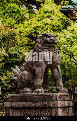 Wächter Komainu-Löwenhund Steinstatue auf Sockel umgeben von üppigem Grün am Nezu-Schrein, Bunkyō Ward, Tokio, Japan Stockfoto