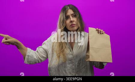 Frau zeigt mit dem Finger und hält die braune Papiertüte vor violettem Studio-Hintergrund, während sie nach links drückt; Frustration. Stockfoto