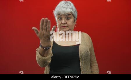 Frau zeigt mit gestreckter Hand und starkem Ausdruck mit dem Finger auf die Kamera, trägt Strickjacke und Tanktop vor rotem Studiohintergrund; beschimpft die Schuld. Stockfoto