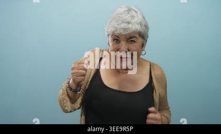 Frau, die Arme hebt und lächelt, mit freiliegenden Schultern und Creolen vor blauem Studiohintergrund, mit Strickjacke und Tanktop; Freude feiern Stockfoto