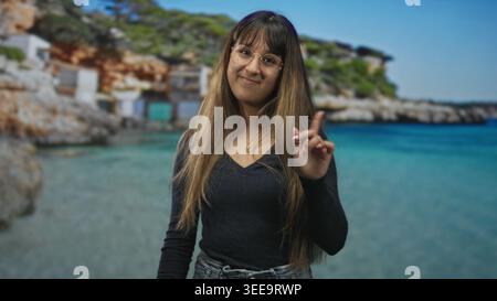 Junge Frau zeigt mit dem Zeigefinger auf die Kamera am Strand, trägt Brille und Denim, lange Haare sichtbar, lächelt sanft; verspieltes Selbstvertrauen. Stockfoto
