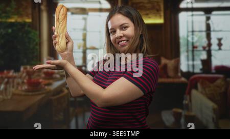 Junge Frau, die ein Baguette mit Handfläche hochhält und Geste im Speisesaal des Hotels in der Nähe von Tisch und Stühlen präsentiert; verspielter Moment. Stockfoto