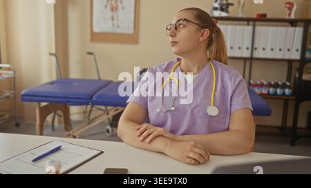 Frau Ärztin in lila Peelings mit gelbem Stethoskop am Schreibtisch, Hände gefaltet neben Klemmbrett und Untersuchungstisch im Gebäude; nachdenkliche Pflege. Stockfoto