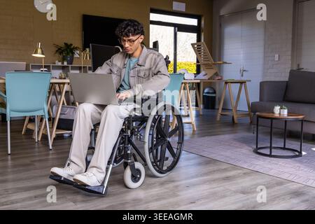 Indischer Mann mit Jeansjacke, im manuellen Rollstuhl sitzend, an einem Laptop in der Gemeinschaftslounge Stockfoto