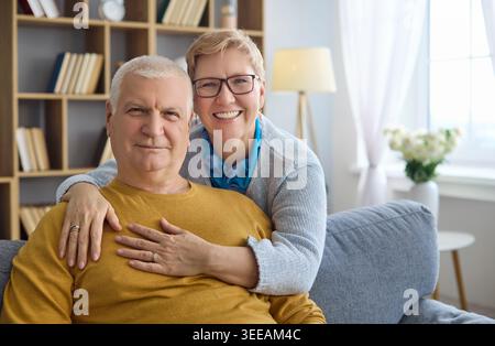 Glückliches Seniorenpaar umarmt sich zu Hause auf dem Sofa, fröhliches Porträt im gemütlichen Wohnzimmer Stockfoto