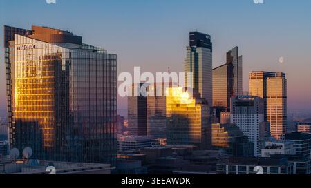 Warschau, Polen - 07. März 2026: Aus der Vogelperspektive auf moderne Wolkenkratzer im Stadtzentrum mit goldenem Sonnenuntergangslicht, das auf Glasfassaden reflektiert, und einem Mond i Stockfoto