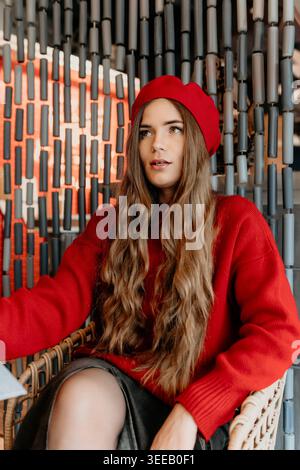 Rote Baskenmütze-Pullover in einem Korbstuhl vor einer modernen strukturierten Wand in einem Café und sieht nachdenklich aus Stockfoto