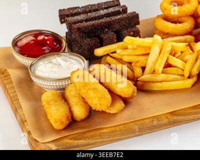 Gemischte gebratene Snacks auf einem Holzbrett, isoliert auf weiß. Goldene Zwiebelringe, Pommes Frites, Käsestäbchen und Roggencroutons mit Ketchup und weißer Sauce. P Stockfoto