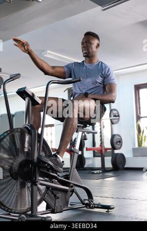 Afroamerikanischer Mann, der im Fitnessstudio auf einem Fächerrad in die Pedale tritt und Gesten treibt, Shorts und Uhr trägt Stockfoto