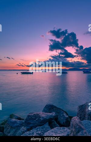 Ruhiger Sonnenuntergang über dem Gardasee mit Booten und Rocky Shore Stockfoto